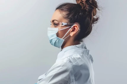 Mujer profesional con mascarilla quirúrgica, gafas de seguridad y bata blanca de laboratorio en vista de perfil.